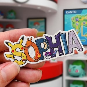 Op de afbeelding: Een gepersonaliseerde sticker of patch met de naam SOPHIA in gestileerde letters. De S is geel met Pikachu-kenmerken, de O is oranje zoals Charizard, de P is donkerblauw, de H is paars, de I is blauw en de A is lichtpaars.