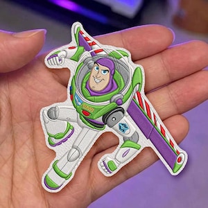 Può includere: Una toppa in tessuto raffigurante il personaggio Buzz Lightyear in una posa dinamica. Il design include una tuta spaziale bianca, verde e viola con una cupola del casco trasparente, bottoni rossi e verdi e una grande ala viola sul retro.