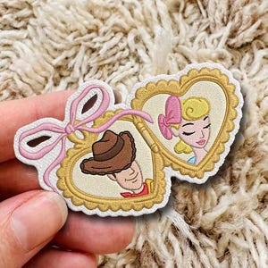 May include: An embroidered patch featuring two connected heart-shaped frames with gold scalloped edges. One frame contains a brown cowboy hat, and the other contains a blonde character with a pink bow. A pink embroidered ribbon ties the two hearts.
