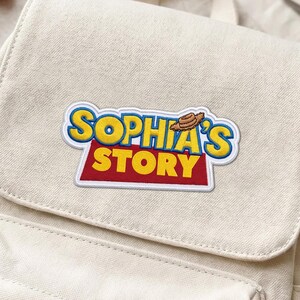 Op de afbeelding: Een crèmekleurige canvas rugzak met een rechthoekige patch op de voorflap met de tekst "SOPHIA'S STORY" in gele en rode letters, geaccentueerd door een klein bruin cowboyhoed-icoontje boven de tekst, met een voorvak en een handvat aan de bovenkant.