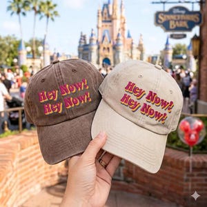 Könnte beinhalten: Zwei Baseballkappen, eine braun und eine beige, jeweils mit dem gestickten Text Hey Now! Hey Now! in einer geschichteten Schriftart in Pink, Gelb und Lila. Die Kappen haben eine strukturierte Krone, einen gebogenen Schirm und einen Knopf oben.
