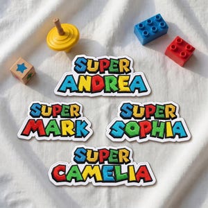 Può includere: Quattro toppe ricamate che riportano la parola SUPER seguita dai nomi ANDREA, MARK, SOPHIA e CAMELIA in uno stile a caratteri cubitali multicolore con spessi bordi neri, disposte su un tessuto bianco con piccoli mattoncini da costruzione.