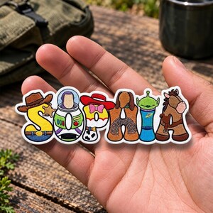 Op de afbeelding: Een geborduurde patch met de naam SOPHIA, waarbij elke letter is vormgegeven naar een ander personage. De S is een cowboyhoed, de O een ruimtepak, de P een cowgirlhoed, de H een speelgoedhond met een veer, de I een alien en de A een paard.