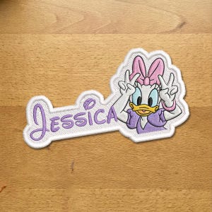 Pode incluir: Um emblema bordado apresentando o nome Jessica em letra cursiva roxa ao lado de um personagem de pato de desenho animado usando um laço rosa, um vestido roxo e fazendo o sinal de paz com as duas mãos, tudo contornado com fio branco.