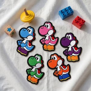 Op de afbeelding: Vijf geborduurde patches met cartoon-dinosaurusfiguren in blauw, roze, paars, groen en oranje. Elk figuur heeft een witte buik, een rood zadel en oranje of blauwe schoenen, geplaatst op een witte stoffen ondergrond.