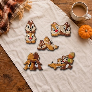 May include: Five embroidered patches featuring the cartoon characters Chip and Dale in various poses, including stacked, hugging, and holding acorns. The patches are rendered in shades of brown, tan, and cream with red noses and black details.