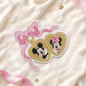 Op de afbeelding: Een geborduurde patch met een goudkleurig hartvormig medaillon met geschulpte randen. Binnenin het medaillon staan portretten van Mickey Mouse en Minnie Mouse. Een grote, lichtroze strik met witte stippen is aan het medaillon bevestigd.