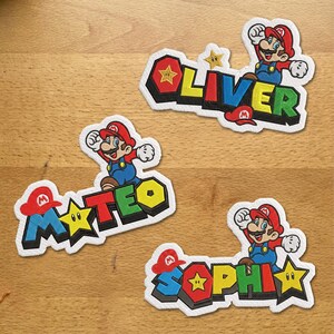 Könnte beinhalten: Drei bestickte Stoffaufnäher mit den Namen Oliver, Mateo und Sophi. Jeder Aufnäher zeigt eine Zeichentrickfigur mit roter Mütze und blauer Latzhose, ergänzt durch ein gelbes Sternsymbol und farbenfrohe Buchstaben in Blockschrift.