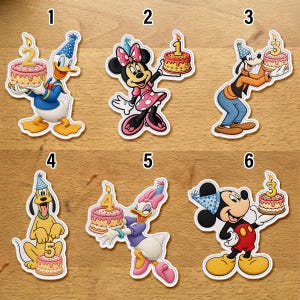 Peut inclure: Six autocollants découpés représentant des personnages Disney avec des gâteaux d'anniversaire. Donald avec un gâteau numéro 2, Minnie avec un gâteau numéro 1, Dingo avec un gâteau numéro 3, Pluto avec un gâteau numéro 5, Daisy avec un gâteau numéro 4 et Mickey avec un gâteau numéro 3.