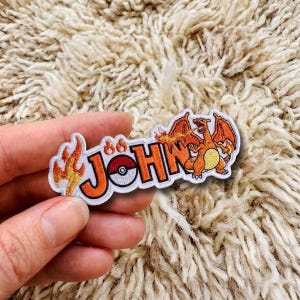 Op de afbeelding: Een geborduurde patch met de naam JOHN in oranje blokletters met een zwarte omlijning. De letter O is vervangen door een rood-witte Poké Ball. De patch bevat oranje vlamaccenten en een afbeelding van de oranje draak genaamd Charizard.