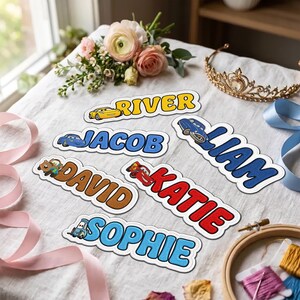 Peut inclure: Six étiquettes de nom personnalisées avec des voitures de course de dessin animé et les noms River, Jacob, Liam, David, Katie et Sophie. Chaque étiquette a une bordure blanche et des lettres colorées. Disposées sur un tissu blanc avec des fleurs.