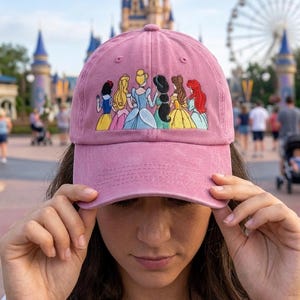 Könnte beinhalten: Eine altrosa Baseballkappe mit einer gestickten Grafik von sechs animierten Prinzessinnen, die in einer Reihe stehen, jede mit unterschiedlichen Haarfarben und Kleidern, darunter Schneewittchen, Aurora, Cinderella, Jasmin, Belle und Arielle.