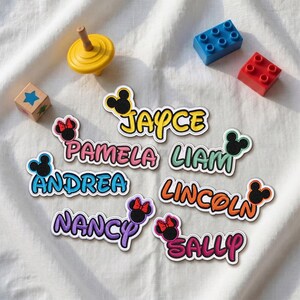 Peut inclure: Sept écussons brodés avec les prénoms JAYCE, PAMELA, LIAM, ANDREA, LINCOLN, NANCY et SALLY. Chaque écusson comporte une icône d'oreilles de souris noire avec un nœud rouge, sur un fond blanc avec une toupie jaune et des blocs de construction.