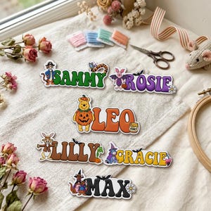 Puede incluir: Cinco etiquetas de nombre personalizadas con personajes de dibujos animados y temas de Halloween. Los nombres SAMMY, ROSIE, LEO, LILLY, GRACIE y MAX están impresos en letras negritas y coloridas, acompañados de ilustraciones temáticas.