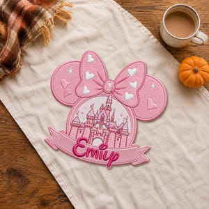 Könnte beinhalten: Ein rosa bestickter Aufnäher mit einer Schloss-Silhouette in einem kreisförmigen Rahmen, gekrönt von einer großen Schleife mit weißen Herzen. Das Design enthält ein Banner am unteren Rand mit dem Namen Emily in dunkelrosa Schreibschrift.