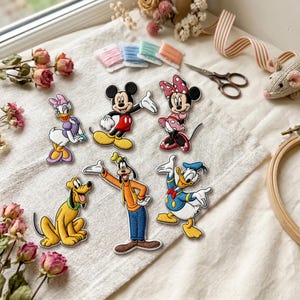 May include: A collection of six embroidered iron-on patches featuring classic cartoon characters including Mickey Mouse, Minnie Mouse, Donald Duck, Daisy Duck, Goofy, and Pluto, arranged on a light-colored fabric surface with sewing supplies.