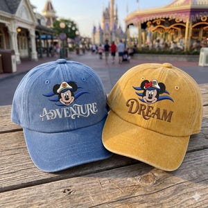 Puede incluir: Una gorra de béisbol azul con un Mickey Mouse bordado con gorra de capitán y la palabra ADVENTURE, y una gorra amarilla con una Minnie Mouse bordada con lazo y la palabra DREAM, ambas con un acabado de tela desgastada.