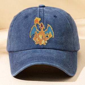 Puede incluir: Una gorra de béisbol azul marino con una textura desgastada y lavada. El panel frontal presenta una colorida ilustración gráfica de tres criaturas naranjas parecidas a dragones, con la más grande escupiendo fuego y dos más pequeñas debajo.