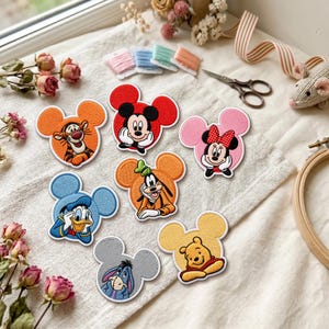 May include: A collection of seven embroidered patches shaped like Mickey Mouse ears, each featuring a different character: Tigger, Mickey Mouse, Minnie Mouse, Donald Duck, Goofy, Eeyore, and Winnie the Pooh, all displayed on a textured fabric surface.