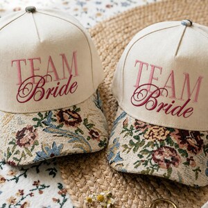 Peut inclure: Deux casquettes de baseball blanc cassé avec le texte brodé "TEAM Bride" en rose et rouge foncé. Les visières sont ornées d'un motif floral tissé dans des tons de vert, rouge, bleu et beige, avec un petit bouton sur le dessus.