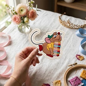 Op de afbeelding: Een geborduurde patch met een bruine cartoonmuis in een geel shirt en groene hoed, die een stapel kleurrijke boeken vasthoudt. De patch heeft een witte gestikte rand en gedetailleerd borduurwerk van het gezicht en de kleding van de muis.
