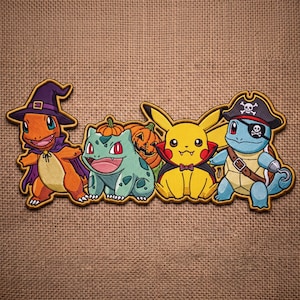 May include: An embroidered patch featuring four characters in costumes: an orange creature in a purple witch hat and cape, a green creature with a pumpkin, a yellow creature in a vampire cape, and a blue creature in a pirate hat with an eyepatch.