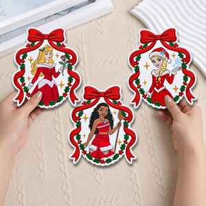 Peut inclure: Trois écussons brodés en forme de boules de Noël, chacun représentant un personnage féminin différent dans une tenue de fête rouge. Les motifs sont encadrés par des rubans rouges, du houx vert et des baies rouges, avec des étoiles dorées.