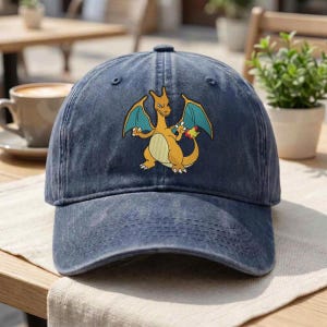 Könnte beinhalten: Eine marineblaue Baseballkappe mit einer mittigen Grafik des orangefarbenen, drachenähnlichen Wesens Glurak, das türkisfarbene Flügel und eine kleine Flamme an der Schwanzspitze hat. Die Kappe besteht aus Stoff mit einer verwaschenen Textur.