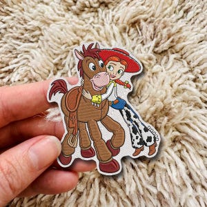 Puede incluir: Un parche bordado que muestra a una vaquera de dibujos animados con cabello rojo, sombrero rojo y pantalones con estampado de vaca abrazando a un caballo marrón con silla de montar. El caballo tiene crines marrones, hocico claro y cascos rojo oscuro.