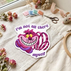 May include: A white embroidered patch featuring a pink and purple striped cat with a wide grin and yellow eyes. The text "I'm not all there" is embroidered in purple above the cat, and the word "myself" is embroidered in purple below the cat.