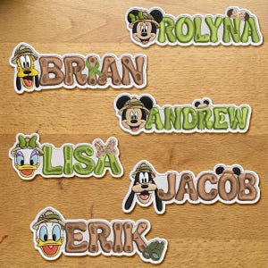 Op de afbeelding: Een collectie van zeven geborduurde naamemblemen met Disney-figuren zoals Mickey, Minnie, Goofy, Donald en Daisy Duck. Elke patch toont een naam in blokletters met een karakterhoofd dat een safarimuts draagt en een verrekijker.