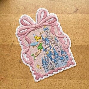 Pode incluir: Um emblema bordado com uma borda festonada rosa claro em forma de etiqueta de presente com um grande laço no topo. O centro retrata uma fada de cabelos loiros e roupas verdes voando ao lado de um castelo com torres e espirais azuis.
