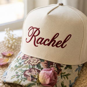 Könnte beinhalten: Eine beige Baseballkappe mit dem Namen Rachel, der in dunkelroter Schreibschrift auf der Vorderseite eingestickt ist. Der Schirm der Kappe ist mit einem gewebten Gobelinstoff bezogen, der ein buntes Blumenmuster mit Rosen und Blättern zeigt.