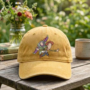 Op de afbeelding: Een mosterdgele baseballpet met een borduursel van twee gezichtsloze stripfiguren, één in een groen en paars ruimtepak en de andere in cowboykleding, geplaatst in het midden van het voorpaneel van de pet.