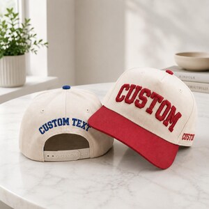Peut inclure: Deux casquettes de baseball blanc cassé avec fermetures snapback réglables. L'une présente une visière rouge et le mot CUSTOM brodé en rouge. L'autre montre l'arrière avec le texte CUSTOM TEXT brodé en bleu et un bouton bleu sur le dessus.
