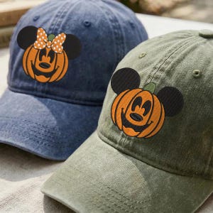 Puede incluir: Dos gorras de béisbol de tela tipo mezclilla, una azul oscuro y otra verde oliva. Cada gorra presenta un parche bordado de una calabaza con forma de cabeza de ratón y orejas negras. La gorra azul tiene un lazo de lunares en la calabaza.