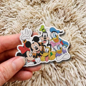 Op de afbeelding: Een gestanste sticker met een groep tekenfilmfiguren, waaronder Mickey Mouse, Minnie Mouse, Goofy, Donald Duck, Daisy Duck en Pluto. De figuren zijn in een groep gerangschikt met heldere kleuren en duidelijke omlijningen op een witte achtergrond.