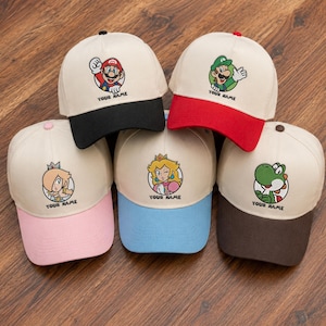 Op de afbeelding: Vijf baseballpetten met gebroken witte kronen en gekleurde kleppen. Elke pet heeft een ronde karakterafbeelding uit de Super Mario-franchise en de tekst YOUR NAME. De kleppen zijn respectievelijk zwart, rood, roze, lichtblauw en donkerbruin.