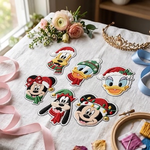May include: A collection of six embroidered fabric patches featuring cartoon characters wearing holiday-themed accessories, including Santa hats, scarves, and festive lights, arranged on a white fabric surface with decorative items nearby.