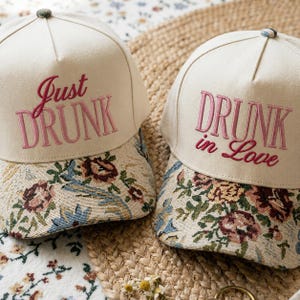 Peut inclure: Deux casquettes de baseball blanc cassé avec des visières en tapisserie florale. La casquette de gauche porte le texte brodé rose Just DRUNK. La casquette de droite porte le texte brodé rose DRUNK in Love. Chaque casquette a un bouton.