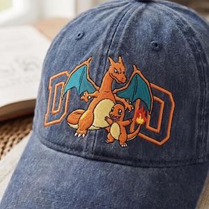 Könnte beinhalten: Eine dunkelblaue Baseballkappe aus Denim mit einer Stickerei der orangefarbenen drachenartigen Kreatur Glurak und der kleineren orangefarbenen Kreatur Glumanda. Das Wort DAD ist in großen orangefarbenen Buchstaben hinter den Figuren gestickt.