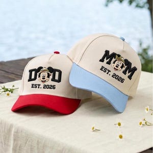 Könnte beinhalten: Zwei Baseballkappen mit cremeweißen Kronen. Die linke Kappe hat einen roten Schirm und zeigt eine Mickey-Maus-Stickerei zwischen den Buchstaben DAD und dem Text EST. 2026. Die rechte Kappe hat einen hellblauen Schirm mit Minnie Maus zwischen MOM und EST. 2026.