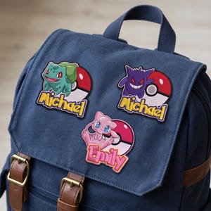 Op de afbeelding: Een marineblauwe canvas rugzak met bruine kunstlederen riemen en gespen. Op de voorklep zijn drie geborduurde patches bevestigd: een groene Bulbasaur met de naam Michael, een paarse Gengar met de naam Michael en een roze Mew met de naam Emily.