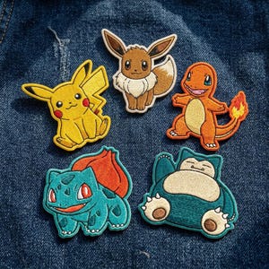 May include: Five embroidered patches featuring cartoon characters: a yellow Pikachu, a brown Eevee, an orange Charmander with a flame tail, a teal Bulbasaur with a bulb on its back, and a large blue Snorlax with a cream-colored belly and closed eyes.