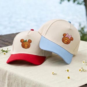Op de afbeelding: Twee baseballpetten met gebroken witte kronen. De linkerpet heeft een rode klep en een geborduurde oranje pompoen met muizenoortjes. De rechterpet heeft een lichtblauwe klep en een geborduurde oranje pompoen met muizenoortjes en een roze strik.