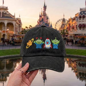 May include: A black baseball cap featuring embroidered designs of a yellow heart-shaped planet with rings, two green three-eyed aliens in blue space suits, and a white and red rocket ship with a heart detail on the front panel of the hat.
