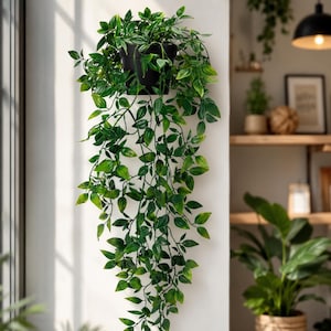 May include: A trailing artificial plant with vibrant green leaves cascading from a simple black pot. The foliage features pointed, oval-shaped leaves in varying shades of green, creating a dense, lush appearance as it hangs downward.