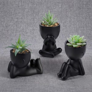 May include: Three matte black ceramic planters shaped like human figures in different poses, each holding a green succulent plant. One figure sits upright, one lies on its side, and one sits with legs extended, all containing small pebbles.