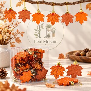May include: A collection of orange maple leaf ornaments, some with a smooth finish and others with a glittery texture. The ornaments feature gold hanging loops and are displayed both hanging from a branch and inside a clear plastic box.