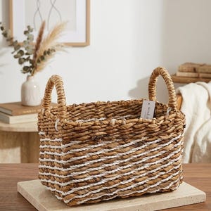 Puede incluir: Una cesta rectangular tejida hecha de fibras de pasto marino marrón natural con un patrón diagonal de cuerda blanca. La cesta cuenta con dos asas tejidas resistentes en la parte superior y una pequeña etiqueta rectangular blanca con texto negro.